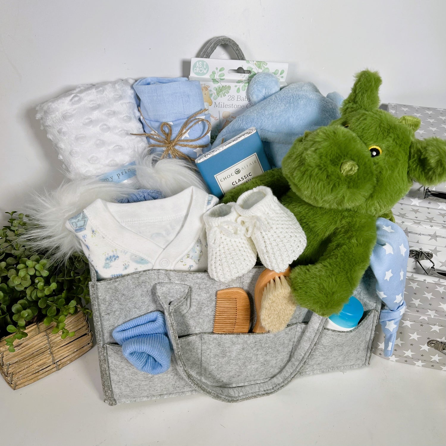 A grey nappy caddy containing a green dragon soft baby toy, a blue and white baby sleepsuit, a blue and white baby pompom hat, a soft white baby blankets, a blue baby dressing gown, a Ziggle baby knot hat and Ziggle baby bib in blue and white stars.